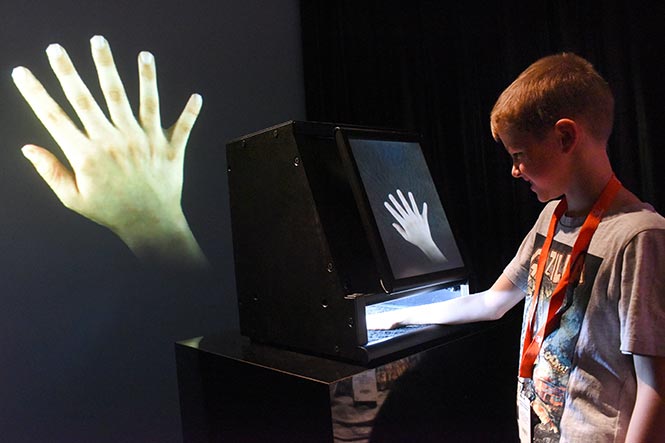 A boy putting his hand inside an experiment box displaying extra fingers on a digital screen