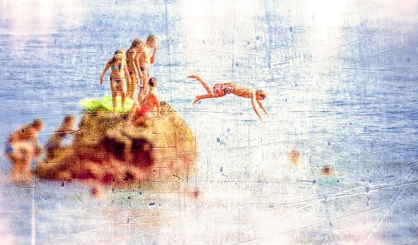 A group of teenagers gathered on a very small rock in the ocean with one jumping into the water